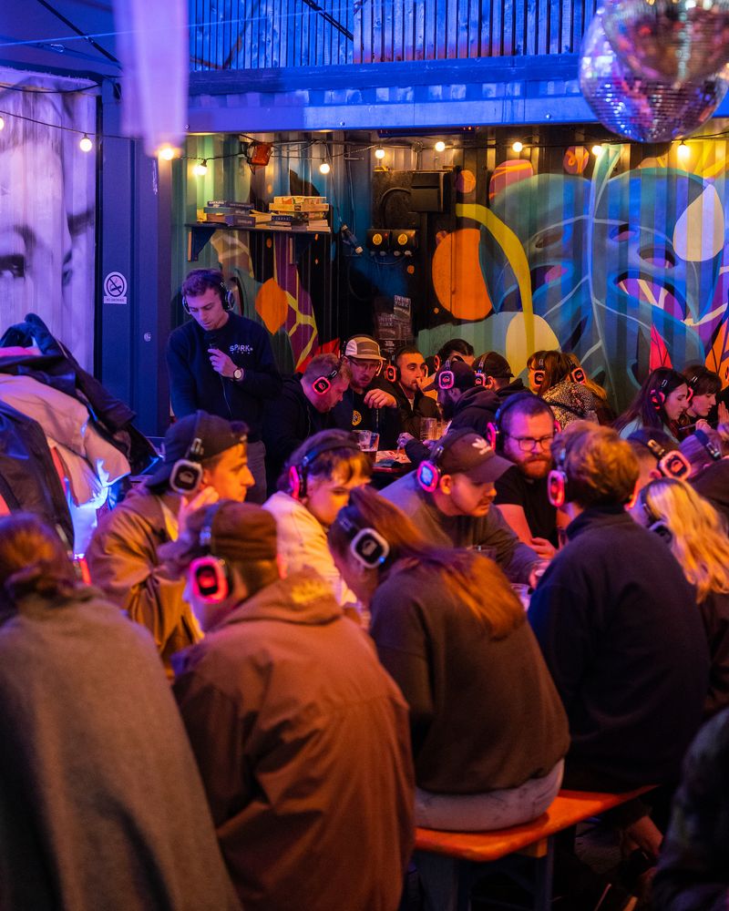 Groups of people wearing headphones whilst co-founder Sam reads quiz questions into a microphone