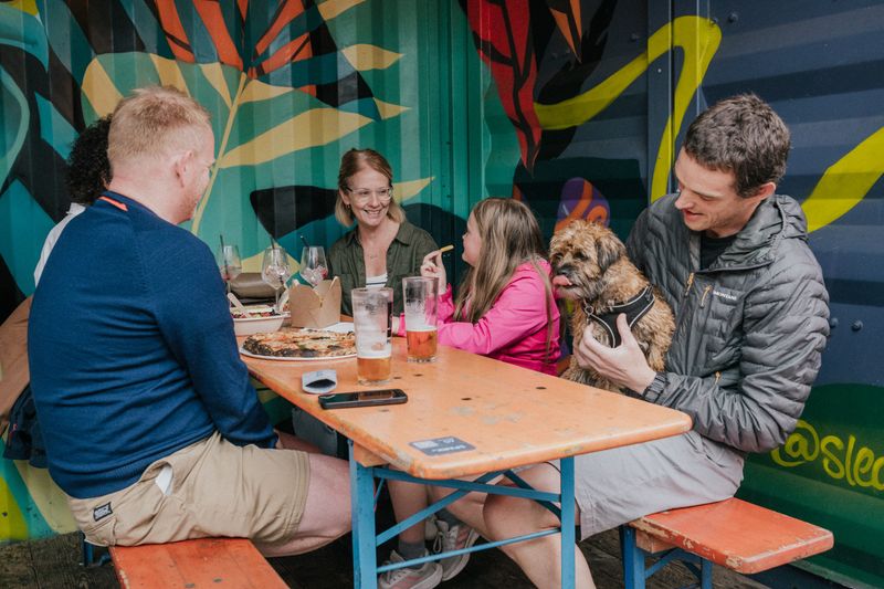 A group of adults and a young girl tuck into food and drink whilst a dog licks their lips whilst sat at the table