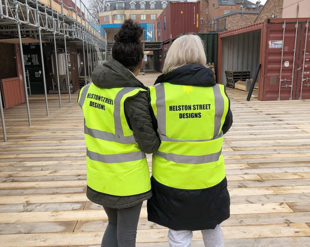 Two women in high-vis jackets with the text "Helston Street Designs" on the back