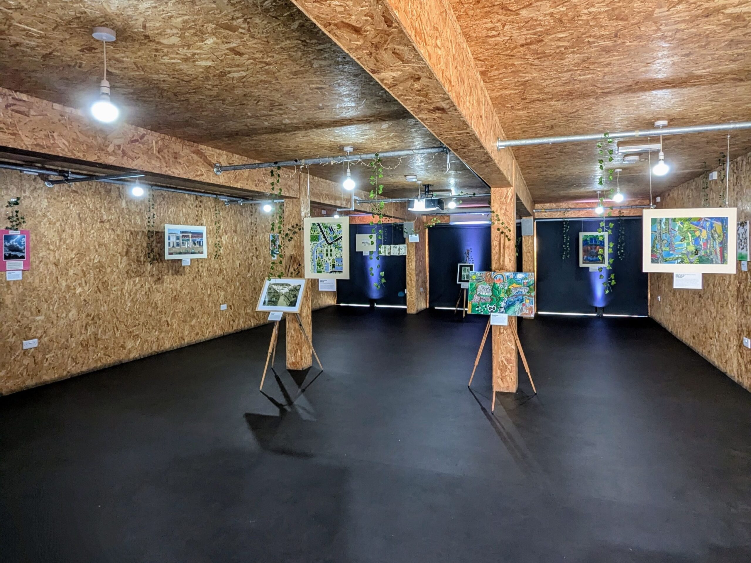 Exhibition room featuring artworks suspended from the ceiling and displayed on easels.