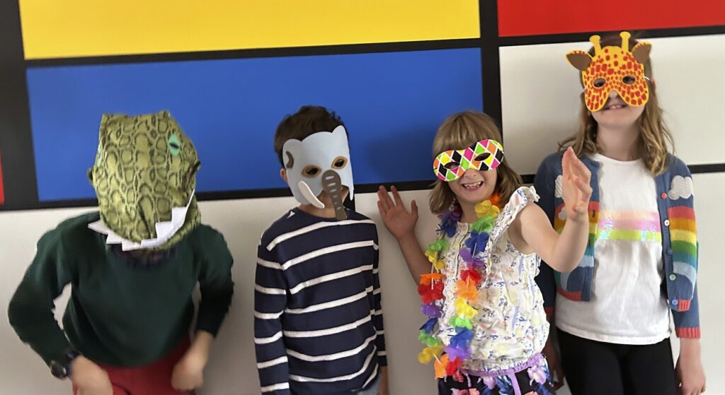 A photo of four children, all wearing masks. One is a dinosaur, one is an elephant, one is wearing a colourful mask, and the other is a giraffe.